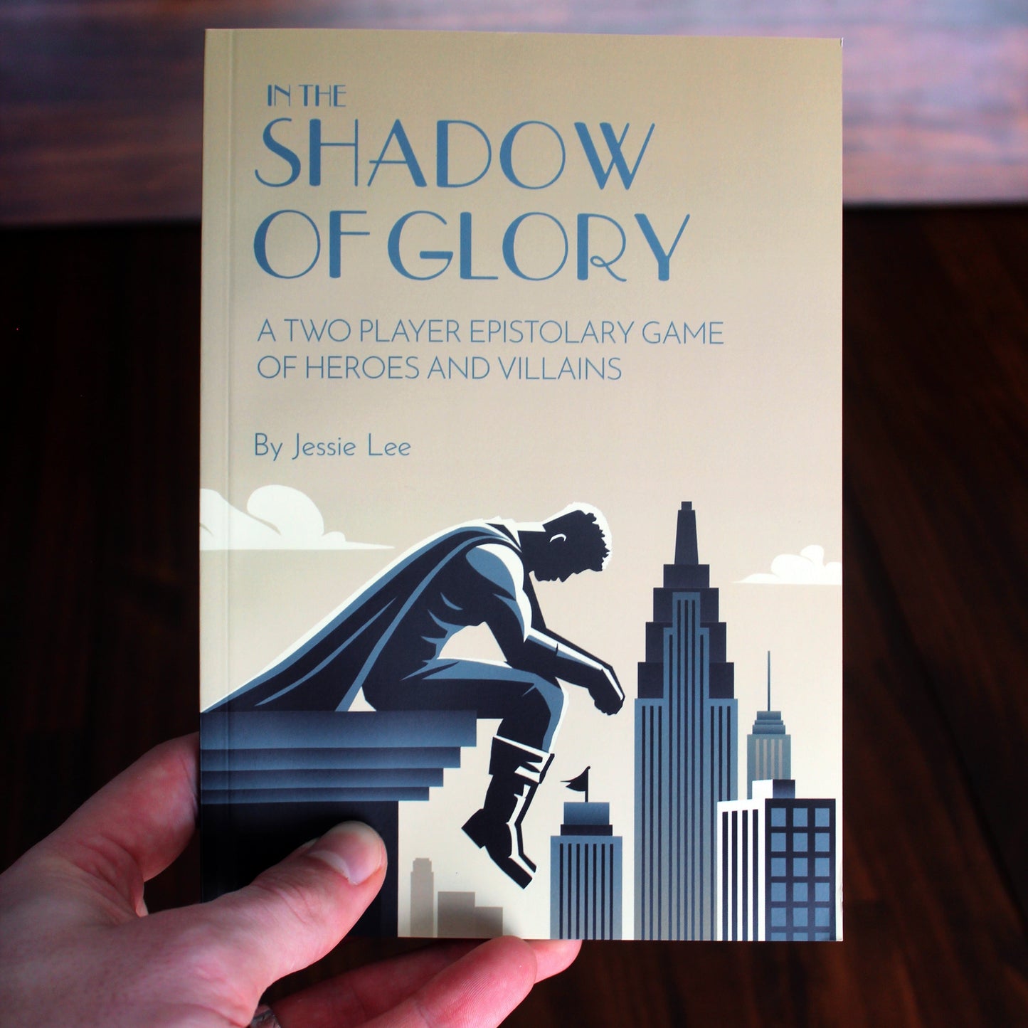 Book titled 'In the Shadow of Glory' held by a hand with a cityscape silhouette on a wooden surface.